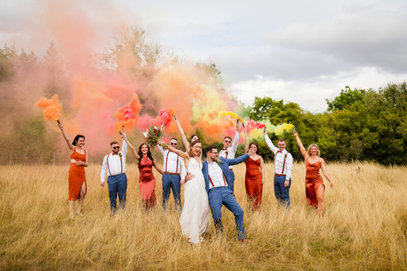 A group of bridesmaids and groomsmen in a field with colored powder captured by a Buckinghamshire wedding photographer.