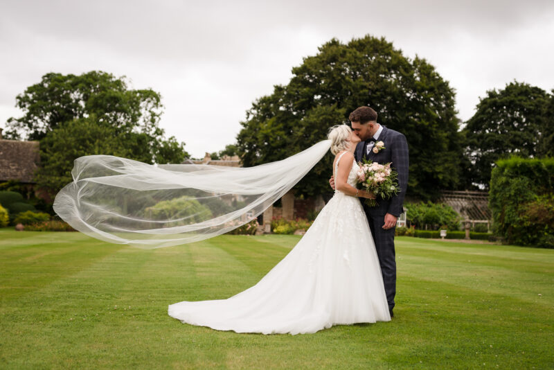 A bride and groom kissing in a field with their veil blowing in the wind.