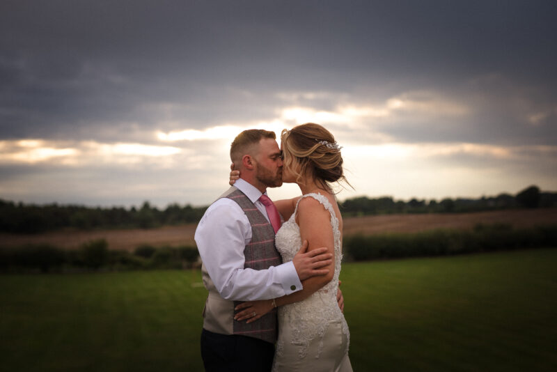 A bride and groom kissing in front of a cloudy sky.